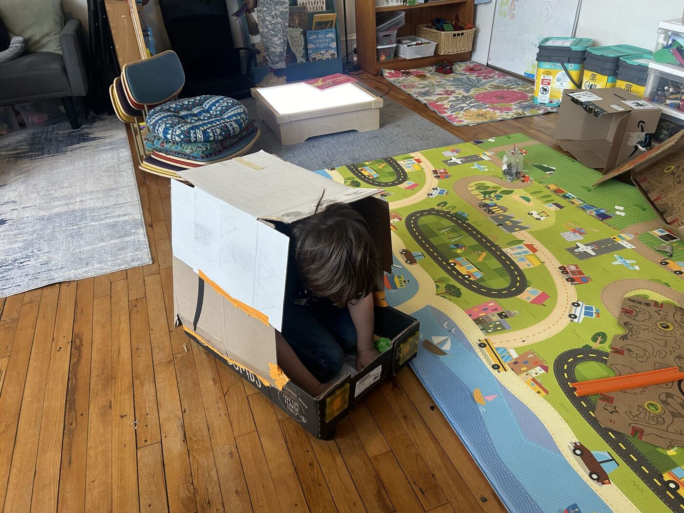 A cardboard box cart on the classroom floor, a learner inside it, head down