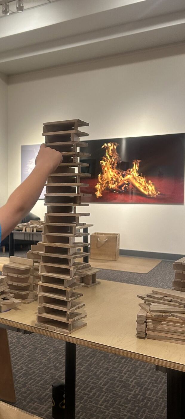 An arm placing the top plank on a tall tower of wooden planks at the science museum
